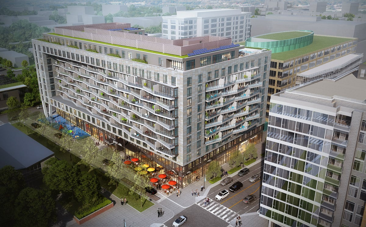 Aerial view of a modern multi-story apartment building with balconies, ground-level shops, outdoor seating with colorful umbrellas, and surrounding city streets and buildings.