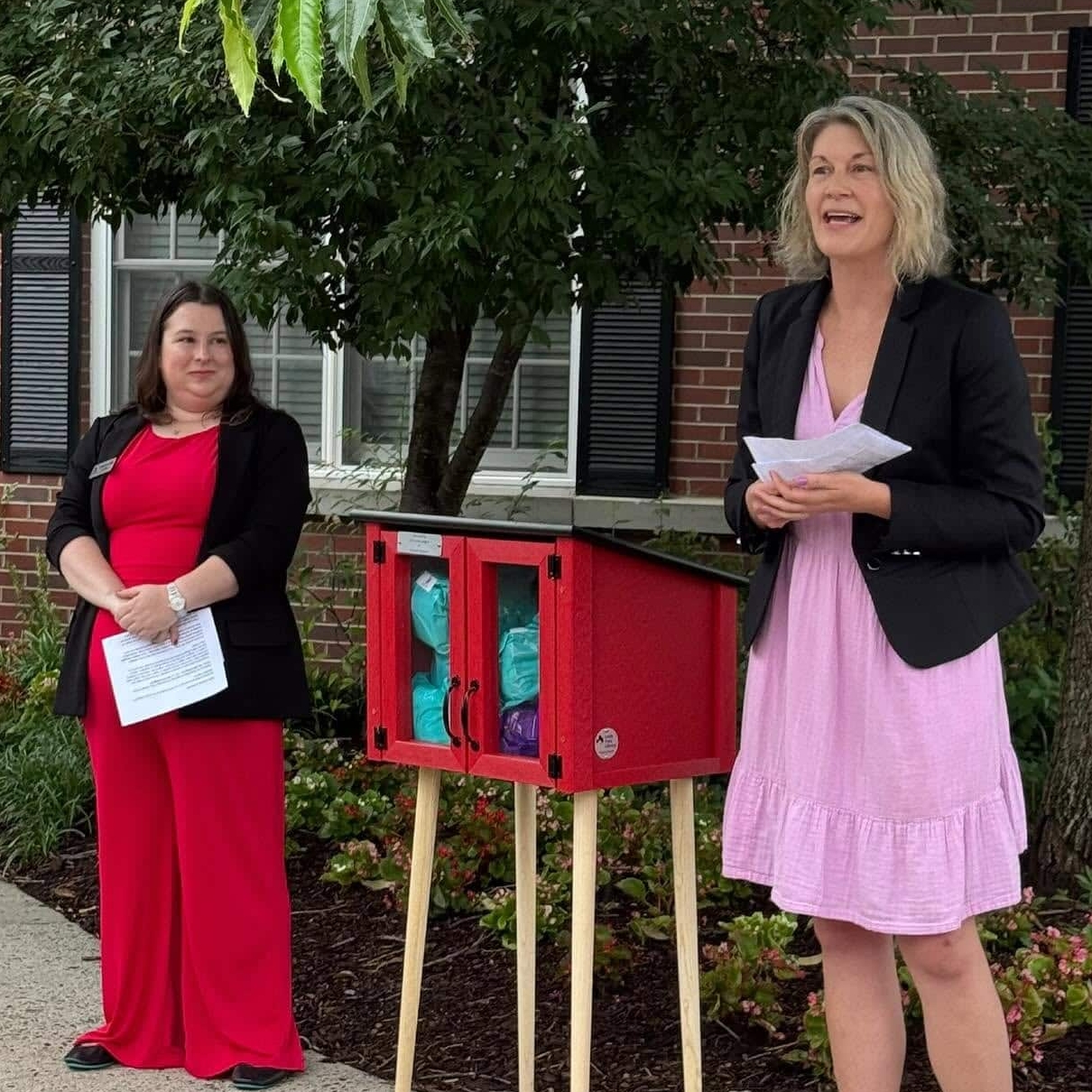 Two women stand outdoors near a small red free library box, as the New Period Pantry opens at Gates of Ballston. One woman in a pink dress and black blazer speaks while the other, in red, holds papers. Trees and a brick building are behind them.