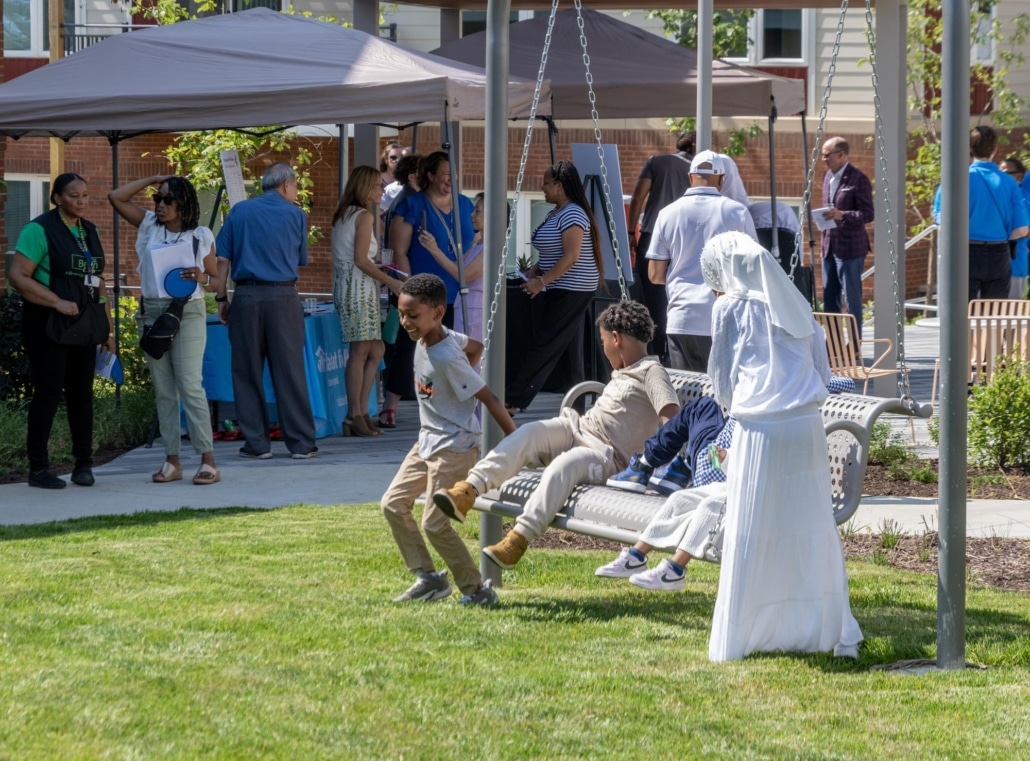 Children play on a swing while a group of adults and other people gather and talk under a canopy at an outdoor event on a sunny day. One child in white stands nearby, watching the others on the swing.