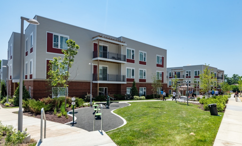 A modern apartment complex with three-story buildings, balconies, landscaped green spaces, walking paths, young trees, and people gathered near a playground under a clear blue sky.