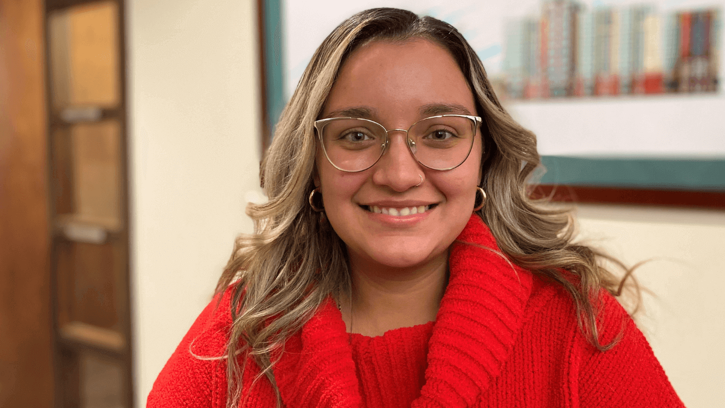 A young woman with glasses and wavy, blonde-highlighted hair smiles while wearing a bright red sweater. She is indoors, with a blurred background featuring a painting and a wooden door.