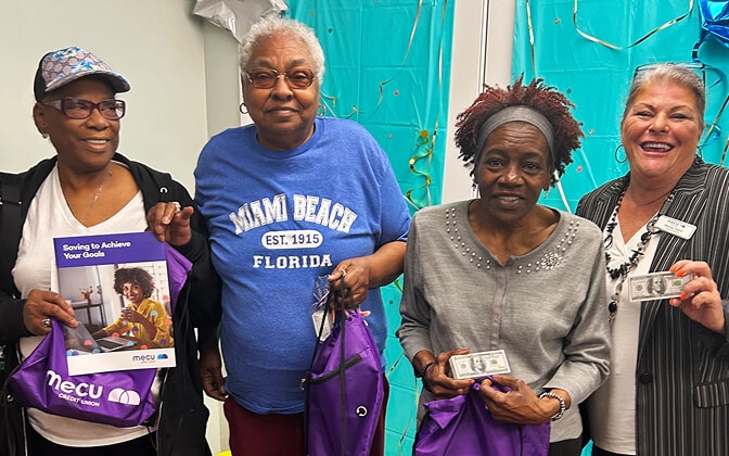 Four smiling older women stand together holding purple tote bags and cash prizes. One woman wears a Miami Beach Florida shirt, another holds a flyer, and festive blue decorations are in the background.