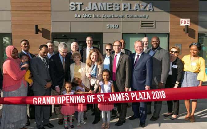 A diverse group of people, including children, stand in front of St. James Plaza holding a large red ribbon that reads, Grand Opening of St. James Plaza 2018. The building’s address, 5140, is visible above the entrance.