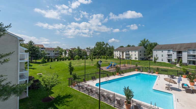Aerial view of an apartment complex featuring a fenced outdoor swimming pool, a playground with slides, green lawns, trees, and several three-story residential buildings under a partly cloudy sky—Building Community With AHC Welcome Baskets.