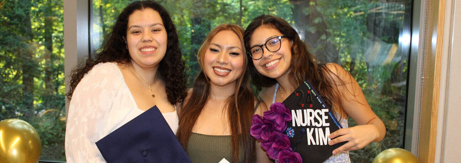 Three young women stand together and smile at the camera, holding graduation caps and diplomas. One cap reads NURSE KING. Green trees, gold balloons, and signs celebrating Affordable Homes & Communities are visible in the background.