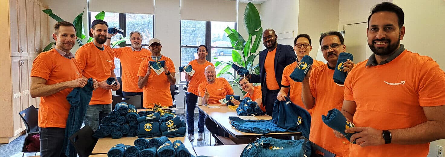 A group of people wearing matching orange t-shirts stand and sit around tables, smiling and folding blue t-shirts in a bright room with large windows and green plants, supporting Affordable Homes & Communities.