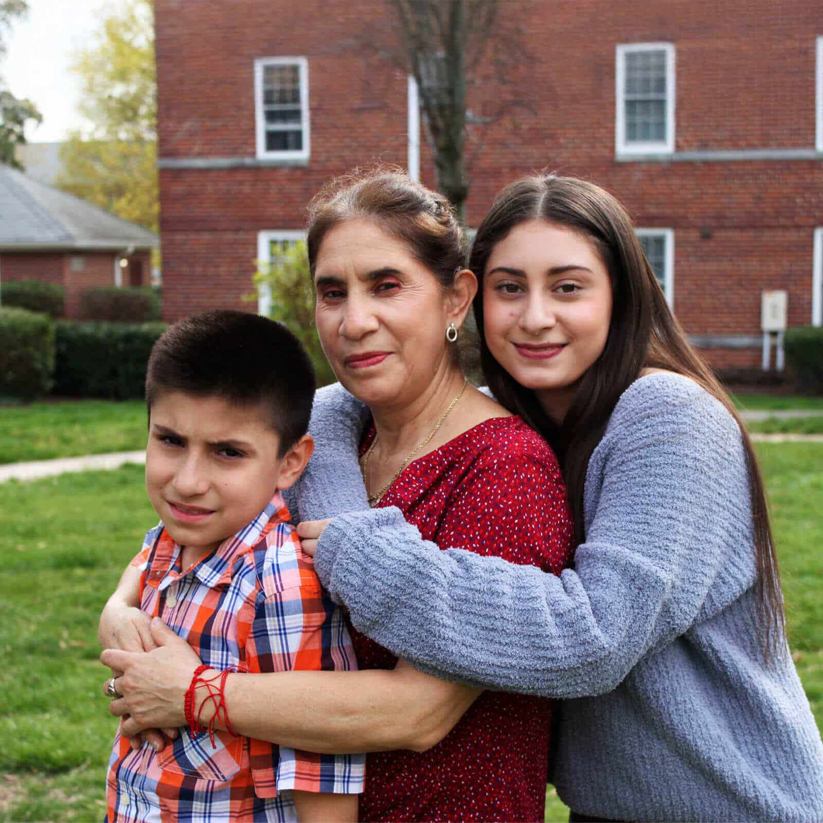 A woman in a red blouse stands outside Affordable Homes & Communities with a young boy in a plaid shirt and a teenage girl in a gray sweater hugging her, all smiling in front of the brick building.