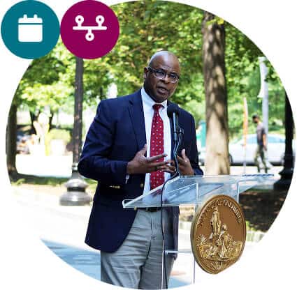 A man in a suit and red tie speaks at a clear podium with a gold seal outdoors in a park, highlighting Affordable Homes & Communities. Two circular icons—a calendar and a network—appear in the top left corner. Trees and a walking path are visible behind him.