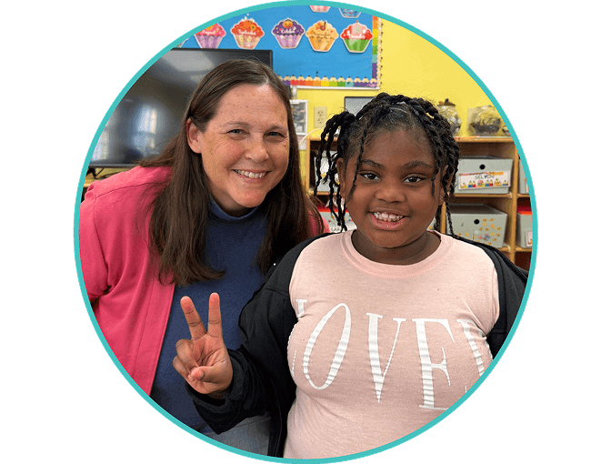 A woman and a young girl smile at the camera in a classroom. The girl holds up a peace sign, and colorful art is displayed on the wall behind them.