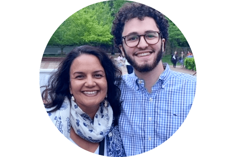 A woman and a man stand close together outdoors, both smiling at the camera. The woman has long dark hair and wears a patterned scarf, while the man has curly hair, glasses, and a checkered shirt. Green trees are in the background.