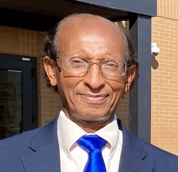 An older man with glasses, short curly hair, and a blue tie smiles while standing outside in sunlight in front of a brick building.