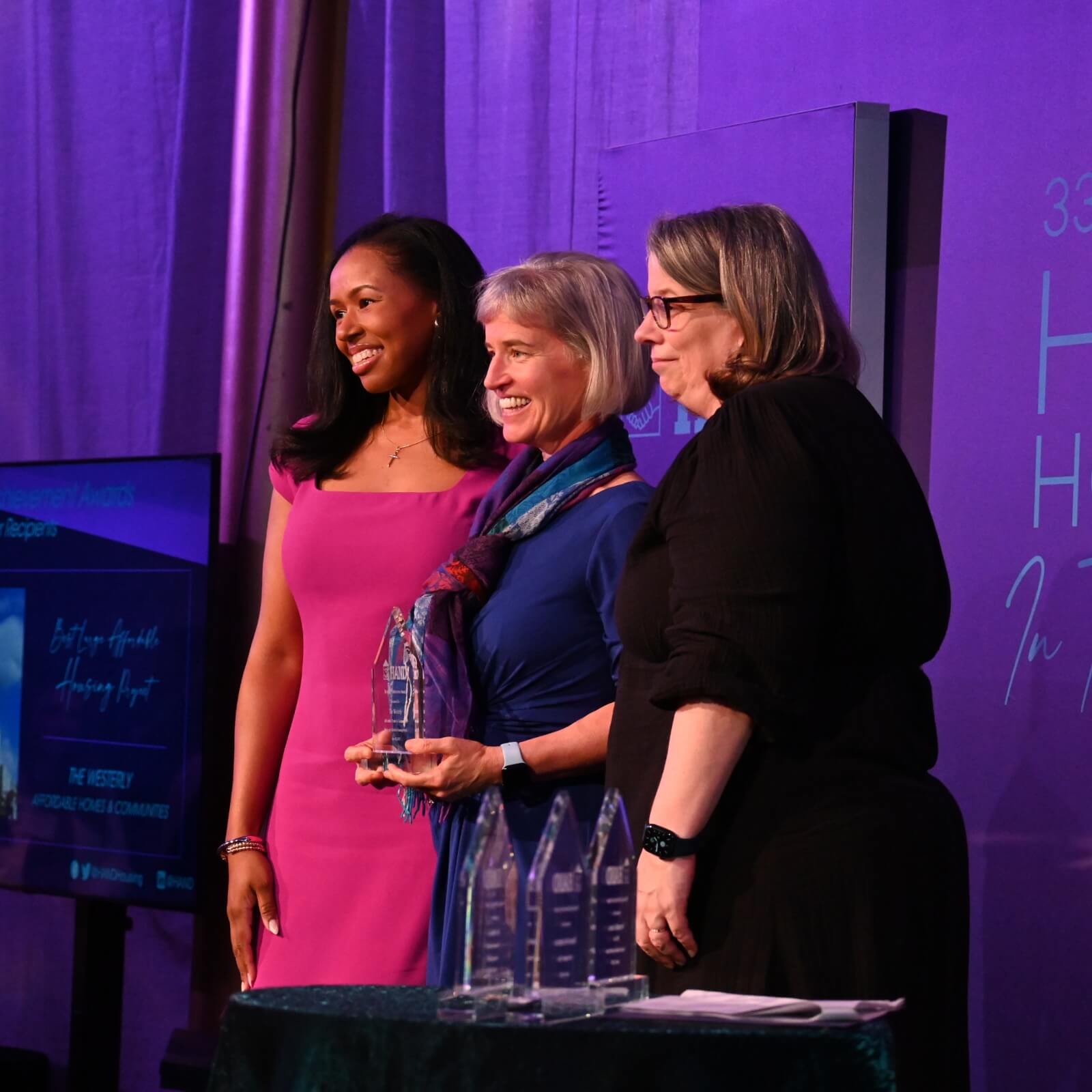 Three women stand together onstage at an affordable housing awards event. The woman in the center holds a glass award, all are smiling. A purple banner is displayed behind them, with several more awards on a table in front.