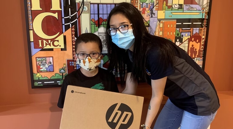A young boy and a woman, both wearing face masks, stand indoors in front of a colorful mural. The boy holds a large HP Chromebook box, highlighting one of the 9 Ways AHC Boosts Economic Mobility in their community.