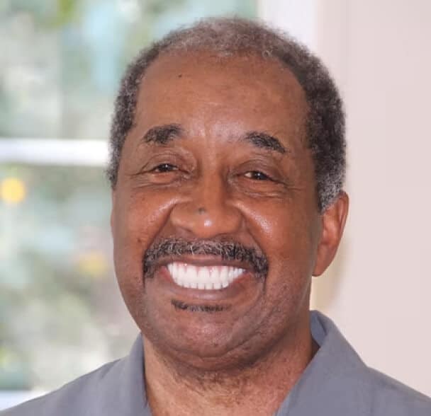 An older man with short curly hair and a mustache smiles widely, showing his teeth. He is wearing a gray collared shirt and is indoors, with a blurred window and greenery in the background.