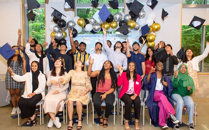 A diverse group of graduates, some seated and some standing, joyfully throw their caps in the air in a decorated room with balloons and a “Congratulations” banner. Everyone is smiling and celebrating.