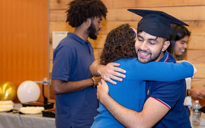 A smiling graduate in a cap hugs a woman at a celebration, while two other people stand nearby, one facing away. Balloons and a cake are visible in the background.