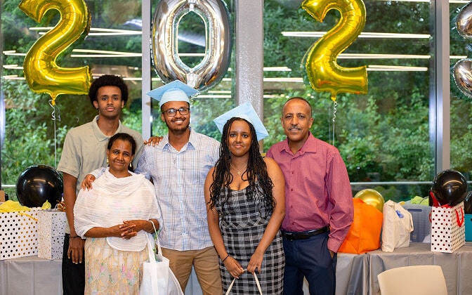 Six people pose together at a graduation celebration with gold balloons forming “2023” behind them. Two people in the center wear blue graduation caps, while others stand around them, smiling. Presents and decorations are on the table nearby.