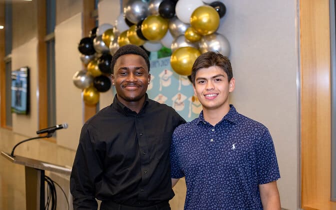 Two young men smile and pose together indoors in front of a backdrop decorated with gold, black, white, and silver balloons. One wears a black shirt; the other wears a navy patterned shirt. A microphone is visible to the side.