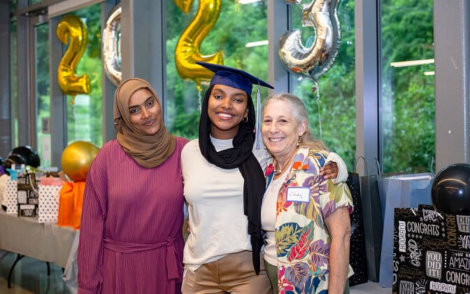 Three women pose together at a graduation celebration. The graduate wears a cap and stands between the other two women. Behind them are 2023 balloons and congratulatory decorations.