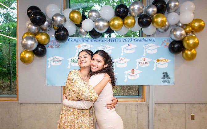 Two young women hug and smile in front of a congratulatory banner for AHC’s 2023 graduates, decorated with gold, black, and white balloons. The banner shows logos of various universities.