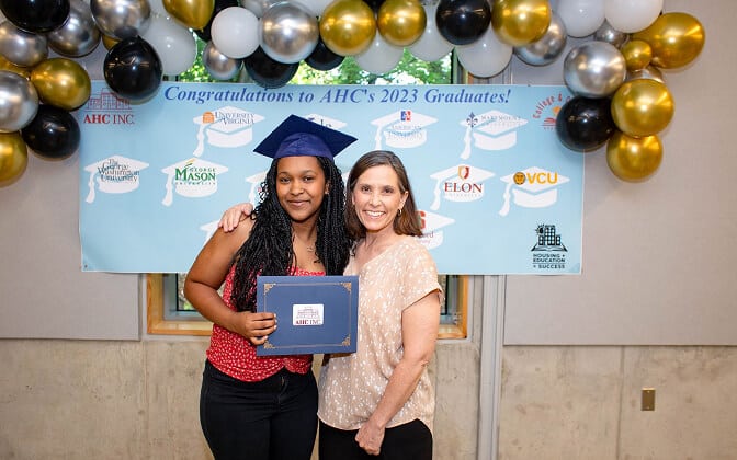 Two women stand smiling in front of a Congratulations to AHC’s 2023 Graduates! banner. One wears a graduation cap and holds a certificate; gold, white, and silver balloons are above them.