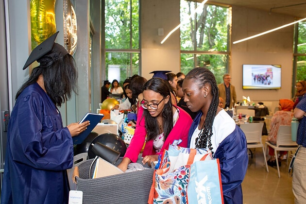 A group of young people, some in graduation gowns, gather around a table with bags and gifts in a bright room. Others are seated in the background. Large windows show green trees outside.
