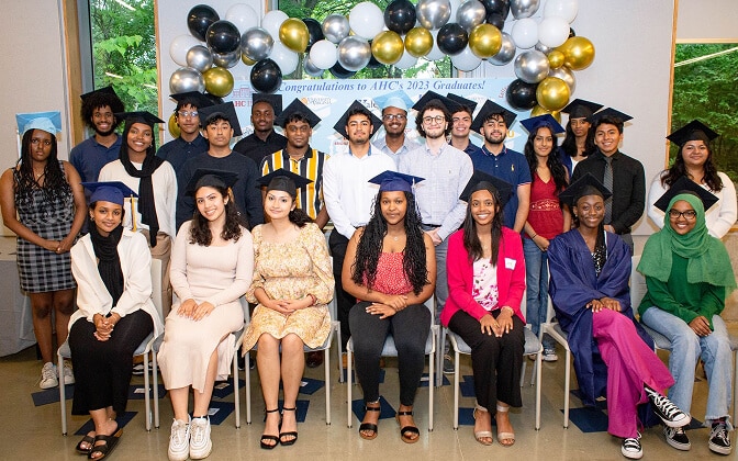 A diverse group of graduating students wearing caps and gowns pose for a photo indoors, seated and standing in two rows, with congratulatory banners and balloon decorations in the background.