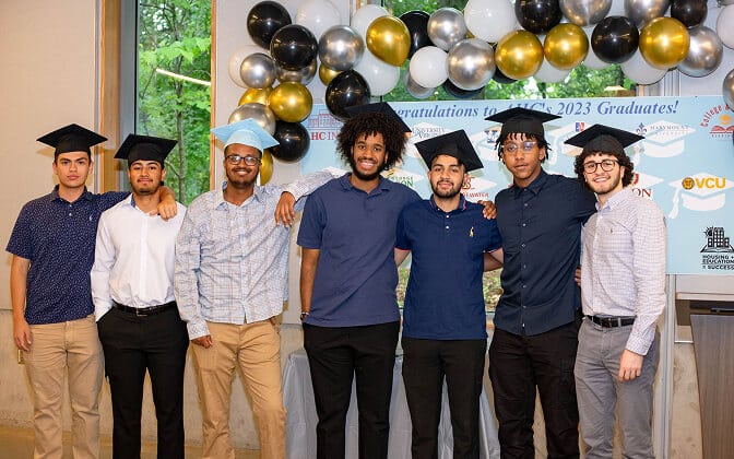 Seven young men wearing graduation caps stand together and smile for a group photo indoors, with a backdrop of balloons and a banner that says “Congratulations to HC 2023 Graduates!”.