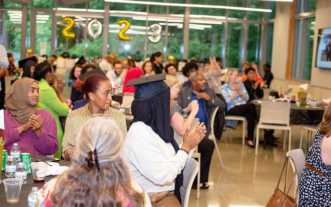 A diverse group of people sits at tables, clapping and facing forward at an indoor event. Graduation-themed “2023” balloons and large windows with greenery outside are visible in the background.