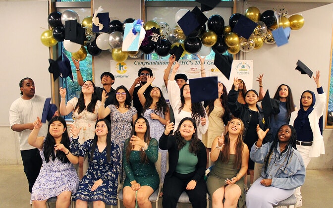A group of diverse students in semi-formal attire celebrate graduation indoors, tossing their caps into the air in front of festive gold, white, and black balloons and a Congratulations banner.