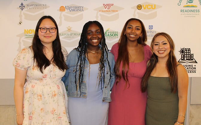 Four young women stand smiling together in front of a backdrop featuring various university logos, including Marymount, UVA, Virginia Tech, VCU, William & Mary, and George Mason.