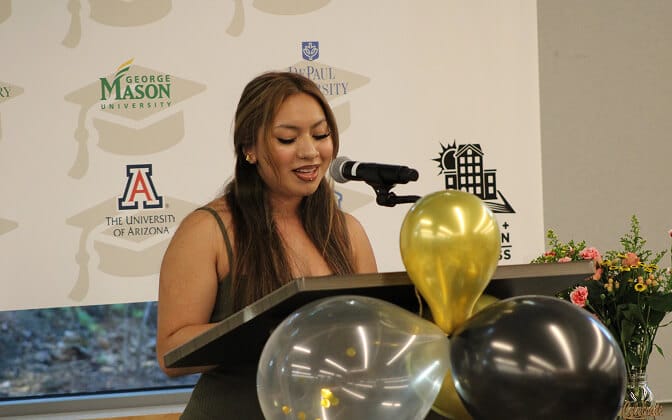 A woman speaks at a podium decorated with black, white, and gold balloons. Behind her is a banner featuring various university logos and a bouquet of flowers is placed nearby.
