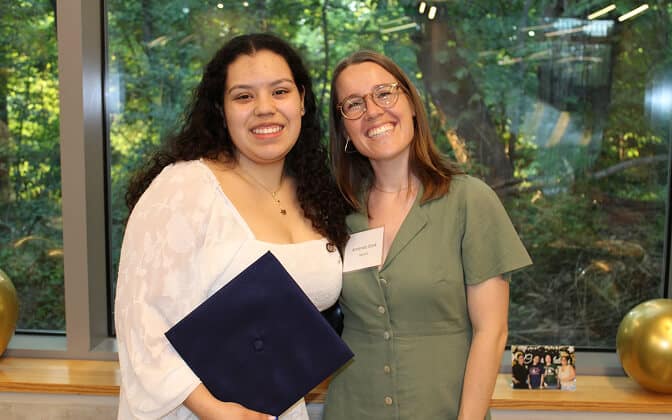Two women smiling and posing together indoors by a large window with trees outside. One woman holds a navy graduation cap and wears white; the other wears green and has a name tag. Gold balloons and a framed photo sit on the windowsill.