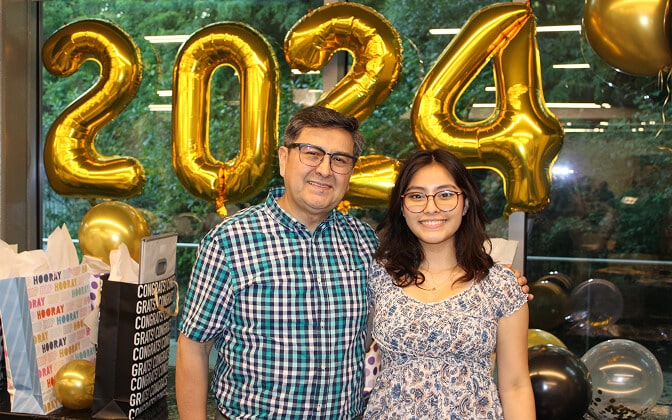 A man and a young woman smile in front of large gold balloons shaped like 2024. Gift bags and balloons are seen beside them, with a window showing green trees in the background.