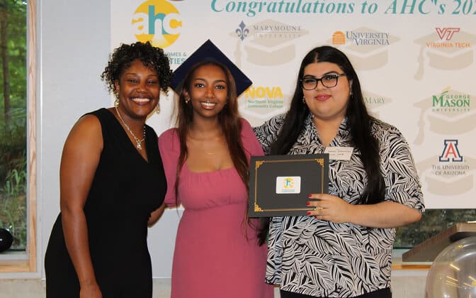 Three women stand together smiling at a graduation event. One woman wears a graduation cap, while another holds a certificate. A congratulatory sign and college logos are visible in the background.