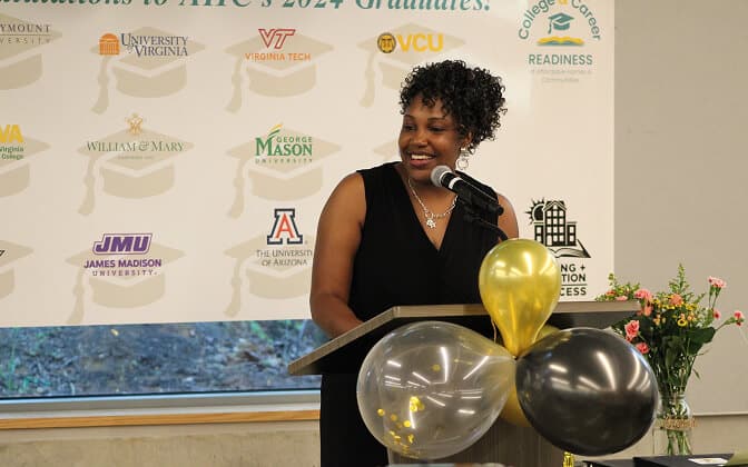 A woman in a black dress speaks at a podium decorated with gold and black balloons. Behind her is a banner displaying various university logos. Flowers are visible on a table to the right.