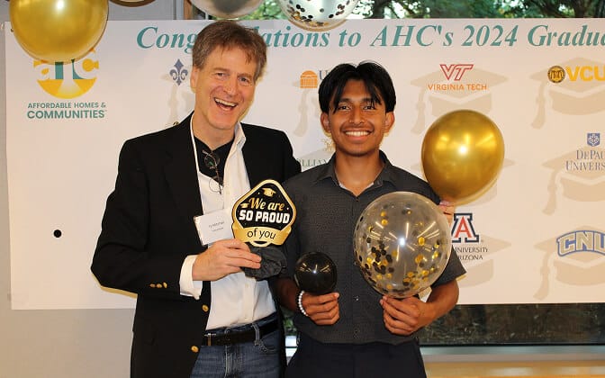 Two men smiling and holding balloons at a graduation celebration. The older man holds a We are SO PROUD of you sign. A congratulatory banner and gold balloons are in the background.