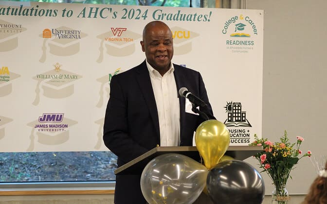 A man in a suit speaks at a podium decorated with black and gold balloons. Behind him is a banner congratulating AHC’s 2024 graduates, featuring various college logos and a vase of flowers on a nearby table.