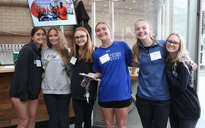 Six young women stand closely together indoors, smiling at the camera. They wear casual clothes and name tags. A TV screen is visible in the background, and the setting appears to be a casual event or gathering.