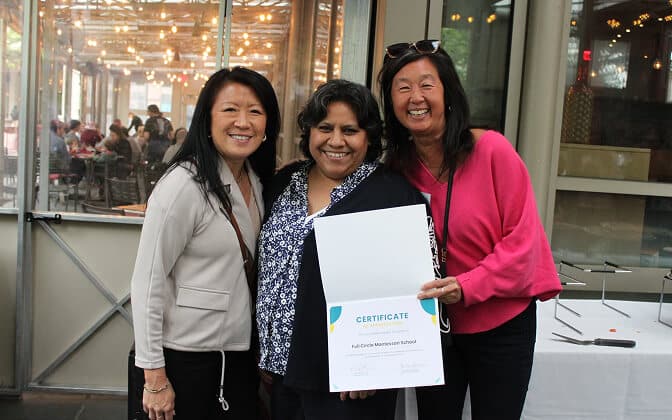 Three women stand together smiling, with one in the center holding an open certificate. They appear to be celebrating an achievement indoors, with a windowed background and string lights visible outside.