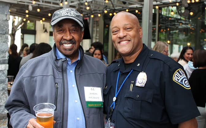 Two men smiling at an outdoor event; one wears a jacket, cap, and name badge, holding a drink, while the other is dressed in a police uniform. String lights and people in the background create a festive atmosphere.