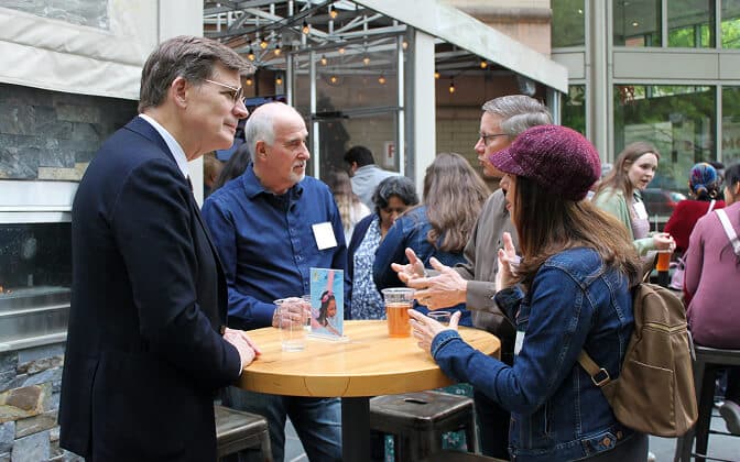 Several people stand around a tall round table at an indoor-outdoor event space, engaged in conversation. Some hold drinks, and everyone appears to be casually dressed and interacting socially.