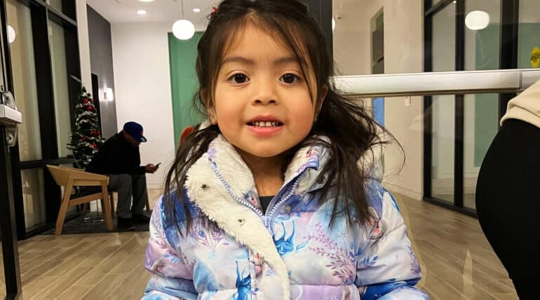 A young girl with long dark hair, wearing a blue and purple winter coat with a fur-lined hood, smiles at the camera indoors. In the background, two adults and a decorated Christmas tree set the scene for Celebrating the Holidays at AHC.