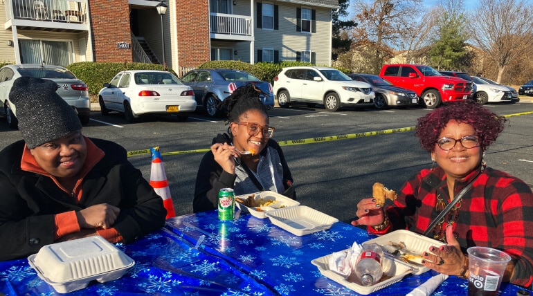 Three people sit at a blue table outdoors, smiling and eating from takeout containers while celebrating the holidays at AHC. Cars and apartment buildings can be seen in the background on this sunny day.