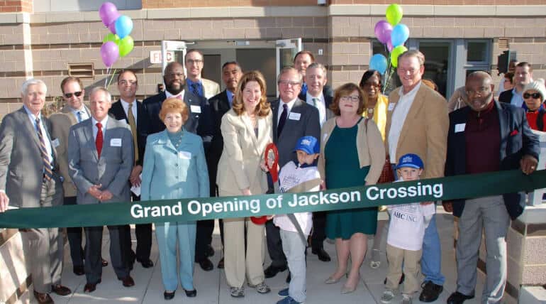 A group of people, including adults and two children, stand behind a green ribbon that reads Grand Opening of Jackson Crossing at an outdoor event, celebrating Building Community With AHC Welcome Baskets amid balloons and a brick building in the background.