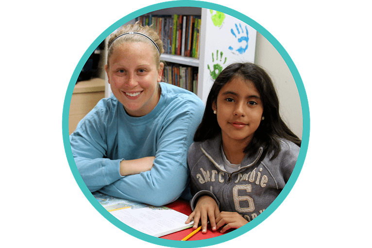 A woman and a young girl sit together at a table with papers and pencils, smiling at the camera. Books and colorful handprints are visible in the background.