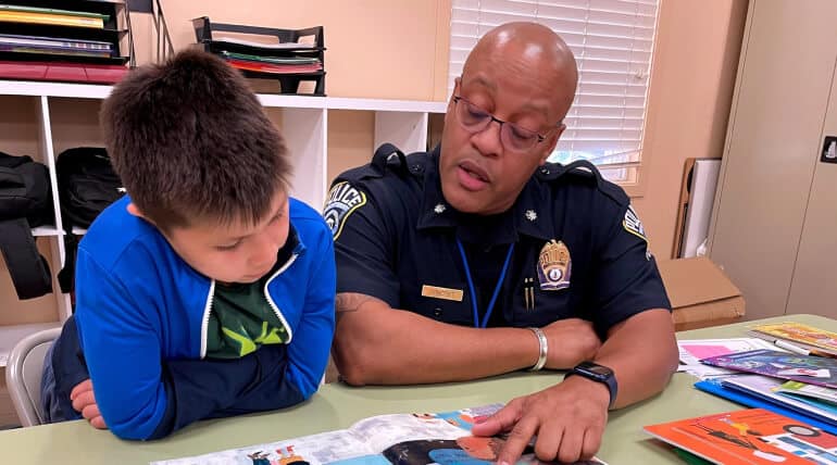 A police officer and a young boy sit at a table, reading a colorful book together. The officer is pointing at the page, while the boy listens attentively. Shelves and stacks of books are visible in the background.