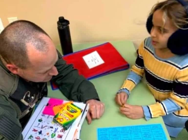 A man and a young girl sit at a table with colorful worksheets, crayons, and folders. The man appears to be assisting the girl, who is wearing earmuffs and a striped sweater.