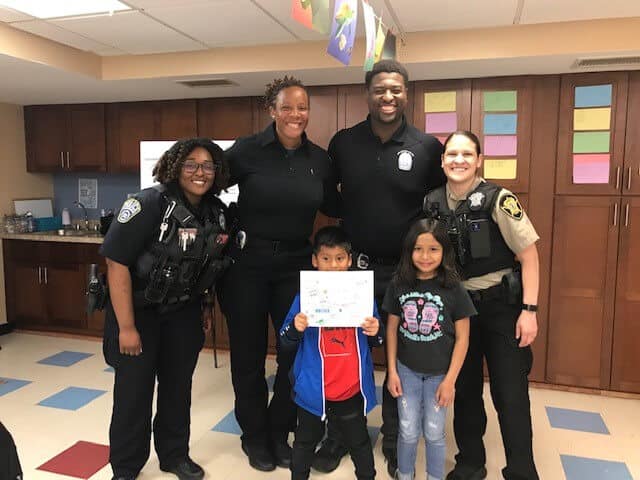 Four smiling police officers pose with two young children in a classroom. The boy is holding a piece of paper with drawings. Colorful papers and flags decorate the background.