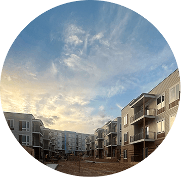 A circular image showing a modern apartment complex with multiple floors, balconies, and a courtyard under a partly cloudy sky at sunset.